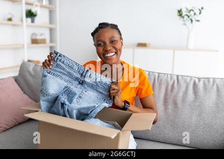 Glückliche schwarze Frau, die Jeans hält, Papppackung entpackt, bestellte Ware erhält, zufrieden mit ihrem Kauf Stockfoto