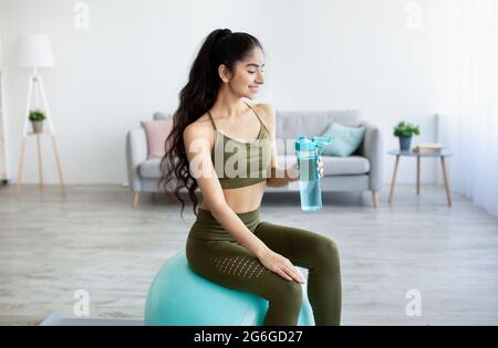Charmante indische Dame in Sportkleidung sitzt auf dem Fitnessball mit einer Flasche klarem Wasser zu Hause Stockfoto