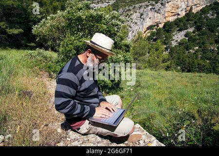 Der Reisende arbeitet fernab auf einem Laptop und genießt dabei den Blick auf die natürliche Berglandschaft im Freien. Mann arbeitet im Freien mit einem Laptop Stockfoto