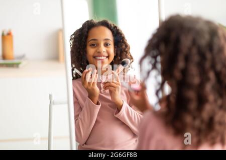 Home Beauty Salon Konzept. Glückliches afroamerikanisches Mädchen, das Make-up macht, Bürsten hält und am Spiegel lächelt Stockfoto