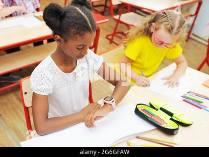 Ein kleines afroamerikanisches Mädchen, das an einem Schreibtisch in einer Grundschule sitzt, sieht vor dem Hintergrund der Schulklasse und auf eine smarte Uhr an ihrer Hand Stockfoto