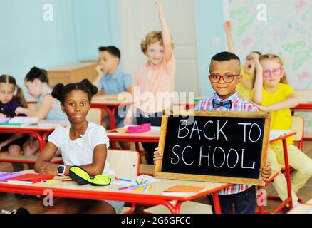 Kleines Kind ein afroamerikanischer Schuljunge mit Brille lächelt und hält vor dem Hintergrund einer Gruppe von Kindern ein Schild mit der Aufschrift Zurück zur Schule Stockfoto