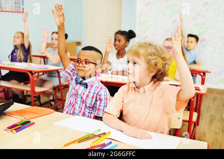 Zwei kleine Schuljungen ziehen die Hand hoch, um vor dem Hintergrund eines Klassenzimmers den Auftrag eines Lehrers in einer Junior School zu beantworten Stockfoto