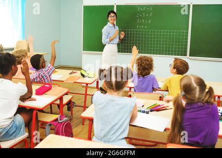 Kinder Vorschulkinder ziehen ihre Hände hoch, um die Frage des Lehrers auf dem Hintergrund der Tafel und des Klassenzimmers in der Grundschule zu beantworten. Stockfoto