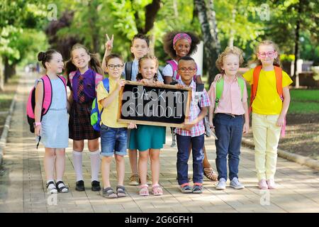 Eine Gruppe von multirassischen Schulkindern in farbenfroher Kleidung, die Schultaschen und Rucksäcke tragen, hält ein Schild mit der Aufschrift „Zurück zur Schule“ gegen die Stockfoto