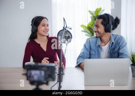 Zwei asiatische Radiomoderatoren, die Podcast im Rundfunkstudio aufzeichnen, eine asiatische Frau, die mit einem Mann online zu Hause am Mikrofon spricht Stockfoto