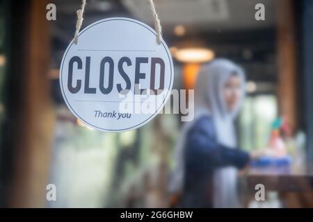 Nahaufnahme Aufhängen an geschlossenem Schild an der Glastür mit Café-Mitarbeitern, muslimischer Angestellter, Frau, die auf dem Tisch putzt, um das Coronavirus zu schützen, oder Covid 19, Stockfoto