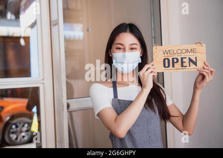 Die Inhaberin des asiatischen Cafés trägt eine Maske, die Frau hält ein Schild, das nach der Stadtsperre geöffnet wird, und ist bereit, zu verkaufen und zu arbeiten Stockfoto