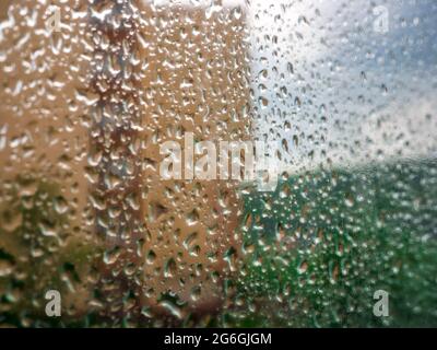 Regentropfen auf das Fensterglas. Verschwommener Hintergrund mit Haus, Grün und bewölktem Himmel - selektiver Fokus Stockfoto