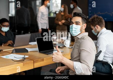 Eine Geschäftsfrau in Maske sitzt am Schreibtisch und zeigt die Daumen nach oben Stockfoto