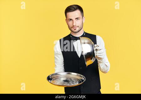 Eleganter junger Kellner, der Cloche über einem leeren Tablett hält, bereit, auf gelbem Hintergrund zu dienen. Restaurant, Horeca, erstklassiges Servicekonzept Stockfoto