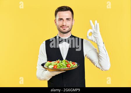 Ein fröhlich lächelnder Kellner hält einen Teller mit gesundem Gemüsesalat, der mit der Hand und den Fingern auf gelbem Hintergrund ein Zeichen tut. Diät, Restaurant, Erfolg Co Stockfoto