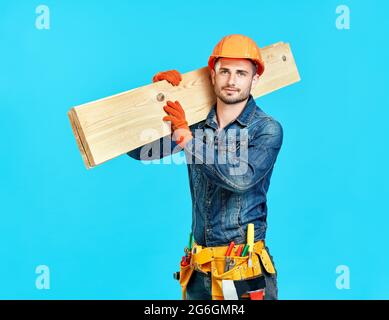 Junger, hübscher Zimmermann hält Holz auf seinen Schultern über blauem Hintergrund und schaut zur Kamera Stockfoto