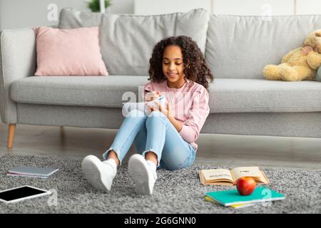 Positive afroamerikanische Teenager Mädchen Hausaufgaben machen, Notizen machen oder Essay schreiben, sitzen im Wohnzimmer Innenraum Stockfoto