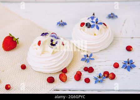 Kleine Baiser-Kuchen, dekoriert mit Schlagsahne, frischen Beeren und essbaren Blüten aus dem echten Leben. Stockfoto