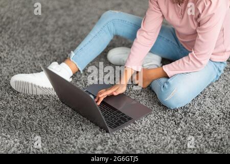 Surfen im Internet. Unkenntlich schwarzes Mädchen sitzt auf Bodenteppich im Wohnzimmer, mit Laptop-Computer, Ernte Stockfoto