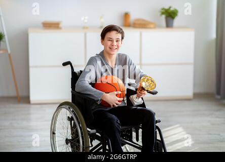 Porträt eines glücklichen behinderten Teenagers im Rollstuhl, der Basketball und einen Preis hält, und lächelt drinnen der Kamera zu Stockfoto