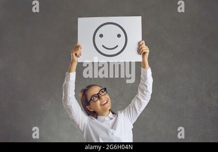 Fröhliche, fröhliche junge Frau, die ein Blatt Papier mit einem Bild eines Smiley-Emojis in der Hand hält Stockfoto