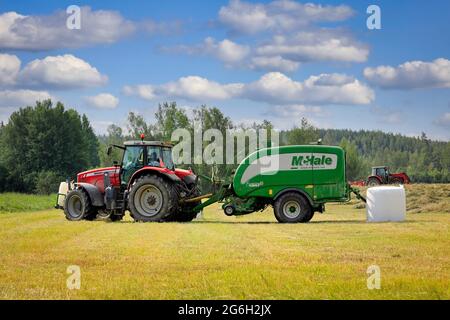 Massey-Ferguson-Traktor im Heufeld mit McHale-Ballenwickler, der im Sommer trockenes Heu erntet. Raasepori, Finnland. 25. Juni 2021. Stockfoto