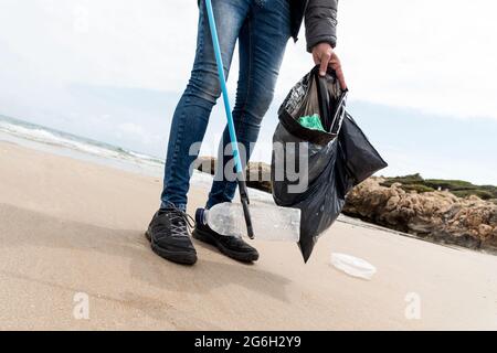 Ein Mann sammelt mit einem Reichweiten-Extender einige Abfälle, wie Dosen, Flaschen oder Taschen, aus dem Sand eines einsamen Strandes, als Aktion, um die Natura zu reinigen Stockfoto