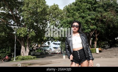 Happy Asian Girl trägt Sonnenbrille Schwarz Lederjacke Schwarz Shorts Weiße Bluse als kausales Model zu Fuß im Park Bangkok Thailand Stockfoto