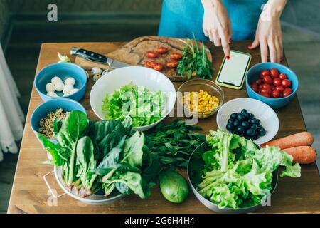 Nahaufnahme eines Küchentischs mit Kräutern und Gemüse. Vegetarische Salatzutaten. Die Hände der Frauen im Telefon Rollen. Stockfoto