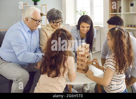 Die glückliche große Familie spielt zu Hause Jenga und wechselt sich ab, indem sie Ziegelsteine aus einem Holzturm herausholt. Stockfoto