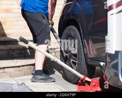 Ein Mechaniker verwendet einen Metallstab auf einem Schraubenschlüssel, um die Radmuttern von einem Wohnmobil-Rad auf einem privaten Antrieb zu entfernen.Wagenheber sichtbar verschwommen im Vordergrund Stockfoto