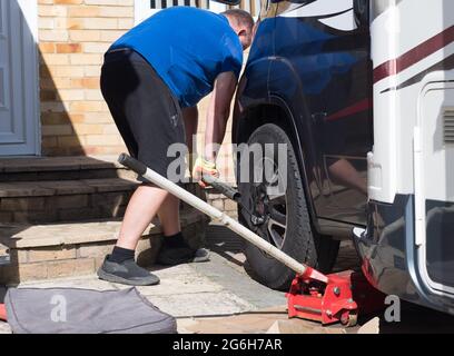 Ein Mechaniker verwendet einen Metallstab auf einem Schraubenschlüssel, um die Radmuttern von einem Wohnmobil-Rad auf einem privaten Antrieb zu entfernen.Wagenheber im Vordergrund sichtbar.Mann Stockfoto