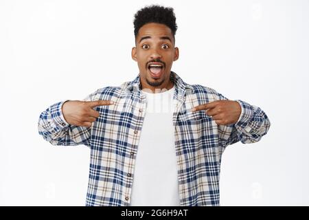 Der verblüffte junge afroamerikanische Hipster macht eine Ankündigung, zeigt mit den Fingern auf den Verkauf, das Logo oder den leeren Raum für Banner, öffnet den Mund vor Ehrfurcht und schaut Stockfoto