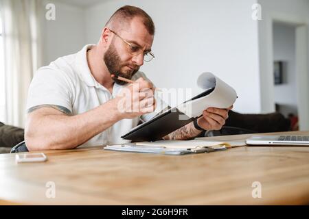 Weißer Mann in einer Brille, der mit Papieren und Laptop arbeitete, während er drinnen am Tisch saß Stockfoto