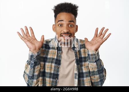 Bild eines afroamerikanischen Mannes, der alarmiert springt, leere Hände hochhebt, Zähne zusammenknallt und unbeholfen und schockiert auf die Kamera starrt und hereinsteht Stockfoto