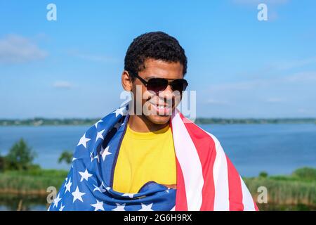 Mann, der die Kamera anschaut und stolz die amerikanische Flagge auf seinen Schultern hält Stockfoto