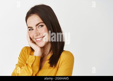 Nahaufnahme einer niedlichen lächelnden Frau, die kokett auf die Kamera schaut, die Wange berührt und errötet, auf weißem Hintergrund im gelben Hemd stehend Stockfoto