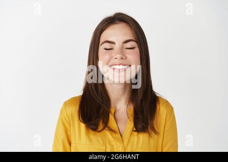 Nahaufnahme einer glücklichen und glücklichen jungen Frau, die mit geschlossenen Augen lächelt, einen Wunsch macht oder träumt, auf weißem Hintergrund stehend Stockfoto