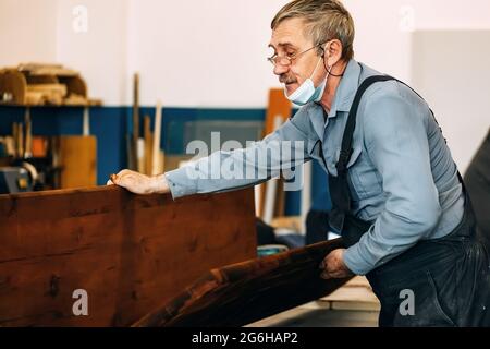 Ein grauhaariger Zimmermann mit Brille arbeitet mit Holz auf einer Werkbank in einer Schreinerei. Ein Rentner in einer medizinischen Maske bei der Arbeit. Beschäftigung und Arbeit ein älterer Mann in Arbeitskleidung Stockfoto