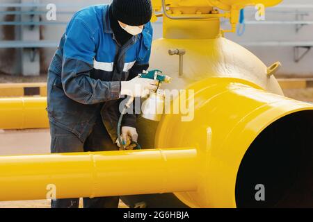 Ein Maler in Arbeitskleidung malt an einem Sommertag ein Absperrventil aus Metall zur Vergasung aus einer Kompressorpistole. Professionelles Lackieren von Teilen. Industrieller Hintergrund. Stockfoto