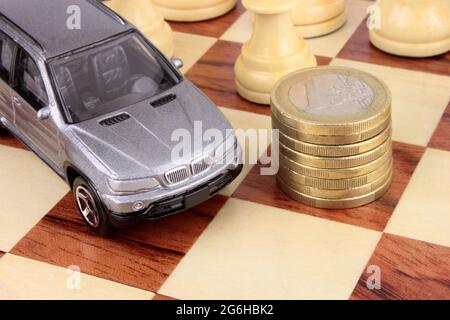 Ein silbergraues Luxusauto auf einem Schachbrett neben einem Stapel Euro-Münzen. Eine Metapher für eine Entscheidung über die Fahrzeugfinanzierung. Stockfoto