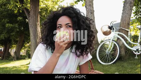 afroamerikanische Frau beißt frischen Apfel in der Nähe Fahrrad im Park Stockfoto