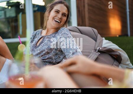 Fröhliche, junge Frau, die sich mit Freunden auf einem Sitzsack entspannt und im Terrassengarten Longdrinks trinkt. Glück und Freundschaft Stockfoto