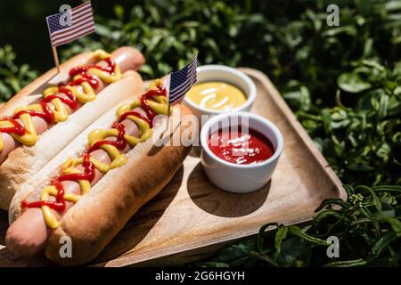 Hot Dogs mit kleinen usa Flaggen in der Nähe von Ketchup und Senf auf Holzschale und grünem Gras Stockfoto