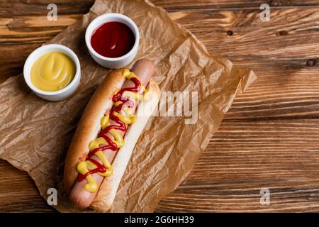 Köstlicher Hot Dog in der Nähe von Schüsseln mit Saucen und zerknittertem Kraftpapier auf Holztisch Stockfoto