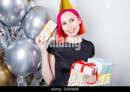 Junge Frau mit rosa Haaren und Partyhut hält Geburtstagsgeschenke und lächelt. In der Nähe Gold- und Silberballons Stockfoto