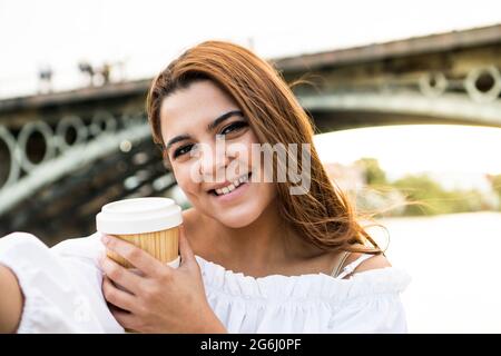 Junge Frau, die ein Selfie beim Kaffee trinkt. Frau, die ein Foto mit dem Fluss und der Brücke im Hintergrund an einem schönen Sommertag in den Spanis macht Stockfoto