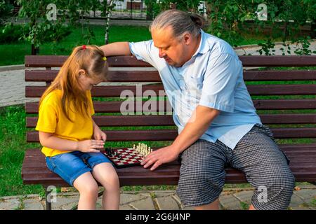 Kleines Mädchen spielt Schach mit Vater im Park auf einer Bank an der frischen Luft. Schachtag. Lehrreiche, lehrreiche Spiele im Freien. Entwicklung von Kindern. Stockfoto