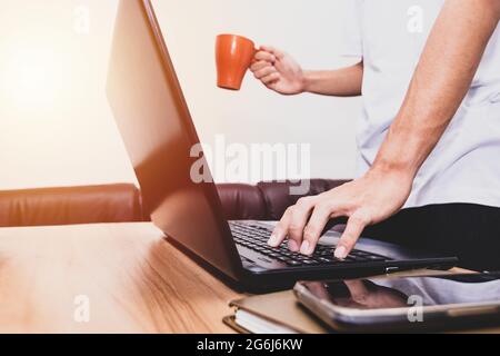 Der Geschäftsmann steht mit einer Kaffeetasse und arbeitet zu Hause an einem Laptop. Technologie, Menschen und Lifestyle-Konzept. Stockfoto