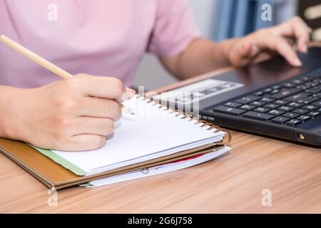 Nahaufnahme einer Frau, die einen Bleistift hält und einen Laptop benutzt, der an ihrem Schreibtisch im Büro arbeitet. Eine Geschäftsfrau schreibt ihre Arbeit in einem Notizschreiben auf Stockfoto