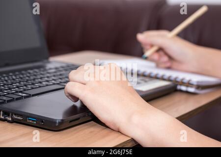 Nahaufnahme einer Frau mit einem Laptop, der an ihrem Schreibtisch im Büro arbeitet. Eine Geschäftsfrau schreibt ihre Arbeit in ein Notizbuch. Arbeiten Sie von zu Hause aus Stockfoto