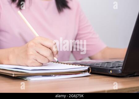 Nahaufnahme einer Frau, die einen Bleistift hält und einen Laptop benutzt, der an ihrem Schreibtisch im Büro arbeitet. Eine Geschäftsfrau schreibt ihre Arbeit in einem Notizschreiben auf Stockfoto