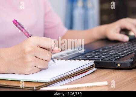 Nahaufnahme einer Frau, die den Stift hält und einen Laptop-Computer benutzt, der an ihrem Schreibtisch im Büro arbeitet. Eine Geschäftsfrau schreibt ihre Arbeit in ein Notizbuch Stockfoto